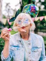 senior stylish woman with gray hair and in blue glasses and jeans jacket blowing bubbles outdoors. Holidays, party, anti age, fun concept photo