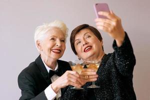 Two beautiful stylish mature senior women drinking wine and making selfie photo. Fun, party, technology, style, celebration concept photo