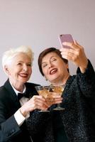 Two beautiful stylish mature senior women drinking wine and making selfie photo. Fun, party, technology, style, celebration concept photo