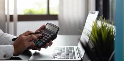 Close up of businessman or accountant hand holding pen working on calculator to calculate business data, accountancy document and laptop computer at office, business concept photo