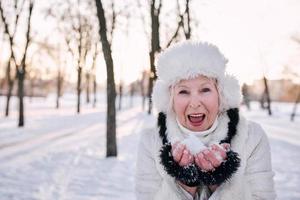 senior woman in white hat and fur coat holding snow in hands in snow forest. Winter, age, season concept photo