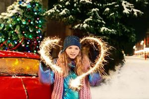 Happy smiling young Caucasian woman in scarf, hat, jacket, mittens with sparkler by the christmas tree outdoor. New year, fun, winter concept photo