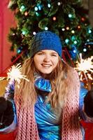 Happy smiling young Caucasian woman in scarf, hat, jacket, mittens with sparkler by the christmas tree outdoor. New year, fun, winter concept photo