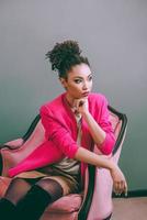 cheerful afro american woman in pink jacket sitting on the pink chair indoor. Fashion, Christmas, new year, happiness, holidays concept photo