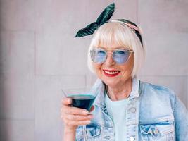 mujer elegante senior con cabello gris y gafas azules y chaqueta de mezclilla con vaso con cóctel azul. alcohol, relax, vacaciones, concepto de jubilación foto