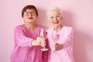 two stylish senior women in pink sweaters drinking rose wine at modern kitchen gossiping. Friendship, talk, gossip, event, relationships, news, family concept photo