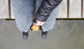 Beautiful legs of a girl in jeans are sitting on a wooden bridge by the lake, holding a yellow maple leaf in their hands. Autumn warm and sunny day. View from above. Autumn concept photo