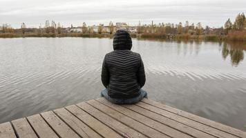 una chica con una chaqueta se sienta en un puente de madera junto al lago en un clima frío de otoño. dipresia o soñar solo. pensando uno a uno con la naturaleza. foto