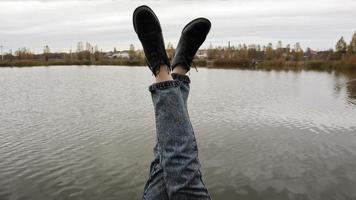 una chica en jeans se sienta junto al lago. una joven descansa junto al río, sentada al borde de un puente de madera. par de piernas relajándose junto a un lago en un muelle de madera. la gente está descansando. foto