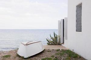 Upside down white boat on the beach, next to a white house and a green plant. Calm sea in the background. Altea photo