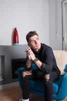 Portrait of a young man sitting in a living room near the fireplace. Dressed in black, looking to his left near the window. photo