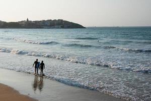 Young couple seen from their back entering the water to surf at Gijon beach at sunset. View of the city in the distance. Asturias, Spain. photo