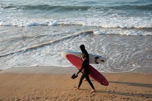 Young couple walking on the beach with their surf boards and wetsuits. Sunset, San Lorenzo beach, Gijon, Asturias, Spain photo