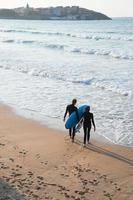 Young couple with wetsuits and surfboards enter the water to surf at San Lorenzo beach, Gijon, Spain. Seen from their back. photo