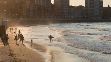 San Lorenzo, Gijon beach and waterfront, at sunset. Unkown people on the sand. A surfer coming out of the sea with his board. Asturias, Spain.