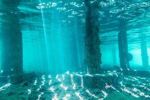 Vista submarina de debajo de un muelle con pilares y luz solar