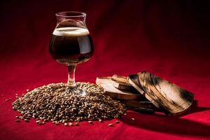 Barril de cerveza oscura envejecida sobre un montón de grano malteado, madera y fondo rojo.