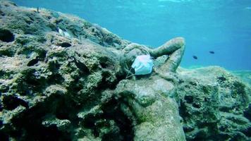 estatua con máscara de medicina en el fondo del mar y varios peces flotando junto a ella como símbolo de la protección contra el covid-19 en los balnearios. video