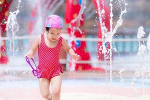Happy little child play with water to cool dancing with fountain at water park in summer or spring.  Girl has fun playing in water fountains. Kid wear red swimsuit. Baby aged 3 years old. photo