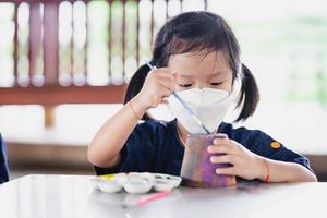 Kid making craft with paintbrush. Little girl wearing surgical white face mask to prevent spread virus and dust pollution PM2.5. photo