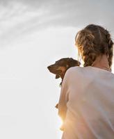 Chica adolescente sosteniendo su perro dachshund en sus brazos al aire libre en la puesta de sol foto