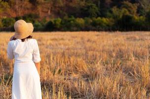 portrait of young woman wearing straw hat and white dress standing in dry rice paddy field at sunset photo