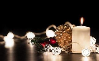 Vela blanca navideña con cono de pino dorado, muérdago y cuerda de luz que brilla intensamente sobre mármol negro foto