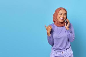 Sonriente mujer asiática sosteniendo un teléfono móvil mientras apunta con el pulgar al espacio de la copia sobre fondo azul. foto