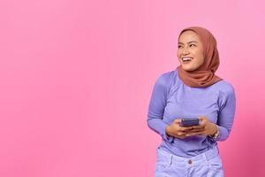 Retrato de mujer asiática joven sonriente sosteniendo el teléfono móvil y mirando a un lado sobre fondo de color rosa foto
