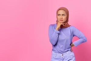 Portrait of pensive young Asian woman touching chin with hand and looking aside on pink background photo