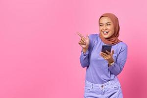 Sonriente mujer asiática sosteniendo un teléfono móvil mientras señala con el dedo en el espacio de la copia sobre fondo rosa