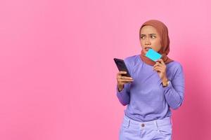 Mujer asiática pensativa sosteniendo teléfono móvil y tarjeta de crédito mientras mira a un lado sobre fondo rosa foto
