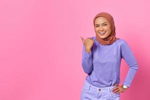 Portrait of smiling young Asian woman pointing a thumb at copy space on pink background photo