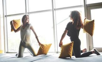 Funny girlfriends play pillows, near large windows. photo