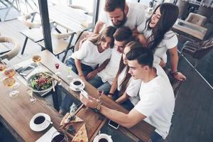 A group of people make a selfie photo in a cafe. The best friends gathered together at a dinner table eating pizza and singing various drinks