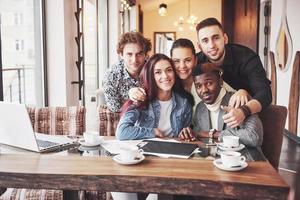Friends having fun at restaurant. Three boys and two girls making selfie and laughing. On foreground boy holding smart phone. All wear casual clothes photo