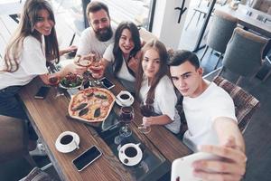 A group of people make a selfie photo in a cafe. The best friends gathered together at a dinner table eating pizza and singing various drinks