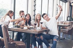 A group of people make a selfie photo in a cafe. The best friends gathered together at a dinner table eating pizza and singing various drinks