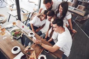 A group of people make a selfie photo in a cafe. The best friends gathered together at a dinner table eating pizza and singing various drinks