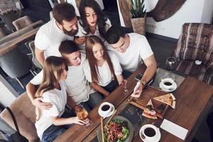 A group of people make a selfie photo in a cafe. The best friends gathered together at a dinner table eating pizza and singing various drinks