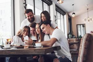 A group of people make a selfie photo in a cafe. The best friends gathered together at a dinner table eating pizza and singing various drinks