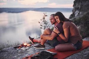 pareja abrazándose con mochila sentado cerca del fuego en la cima de la montaña disfrutando de la vista de la costa, un río o un lago. viajando a lo largo de las montañas y la costa, la libertad y el concepto de estilo de vida activo foto