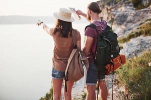 dos turistas masculinos y femeninos con mochilas se paran en la cima del risco y disfrutan del amanecer. viajando montañas y costa, libertad y concepto de estilo de vida activo