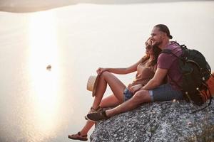 Hugging couple with backpack sitting on top of rock mountain enjoying view coast a river or lake. Traveling along mountains and coast, freedom and active lifestyle concept