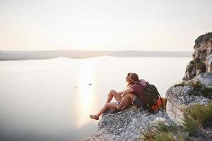 pareja abrazándose con mochila sentado en la cima de la montaña de roca disfrutando de la vista de la costa, un río o un lago. viajando a lo largo de las montañas y la costa, la libertad y el concepto de estilo de vida activo foto