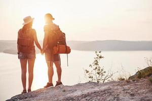 Two tourist male and woman with backpacks stand to the top of the crag and enjoying sunrise. Traveling mountains and coast, freedom and active lifestyle concept