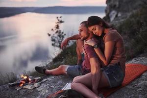 pareja abrazándose con mochila sentado cerca del fuego en la cima de la montaña disfrutando de la vista de la costa, un río o un lago. viajando a lo largo de las montañas y la costa, la libertad y el concepto de estilo de vida activo foto