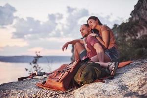 Hugging couple with backpack sitting near the fire on top of mountain enjoying view coast a river or lake. Traveling along mountains and coast, freedom and active lifestyle concept photo