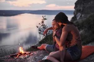 pareja abrazándose con mochila sentado cerca del fuego en la cima de la montaña disfrutando de la vista de la costa, un río o un lago. viajando a lo largo de las montañas y la costa, la libertad y el concepto de estilo de vida activo foto