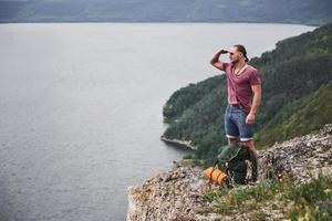 viajero con mochila sentado en la cima de la montaña disfrutando de vistas sobre la superficie del agua. viajando a lo largo de las montañas y la costa, la libertad y el concepto de estilo de vida activo foto